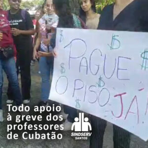 Foto do ato onde uma professora segura um cartaz escrito "PAGUE O PISO JÁ" e uma frase por cima da foto: "Todo apoio à greve dos professores de Cubatão"