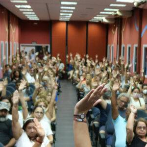 Foto do auditório lotado com os servidores levantando a mão em votação.