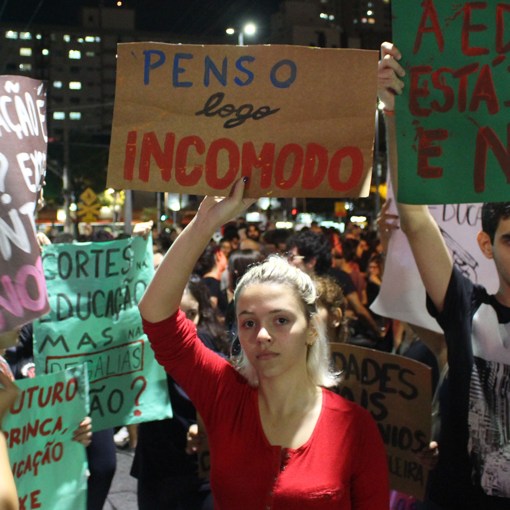 Foto de um ato de rua em Santos com uma estudante erguendo um cartaz escrito "Penso logo incomodo"